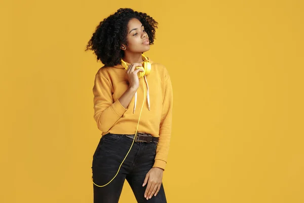 Trendig afrikansk amerikansk ung kvinna klädd i casual kläder kläder lyssna musik bort över gul bakgrund. — Stockfoto