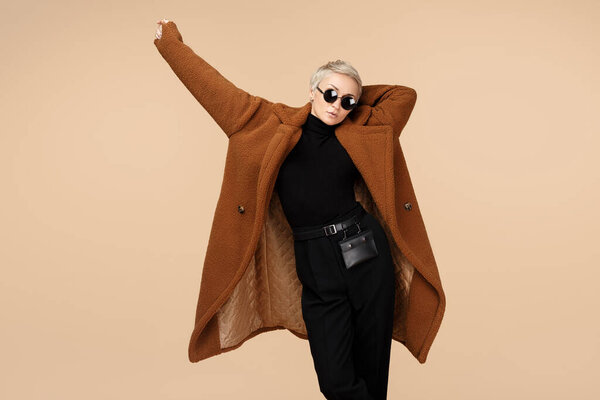 Современная молодая хипстерша с короткими светлыми волосами в пальто и солнцезащитных очках на бежевом фоне
.