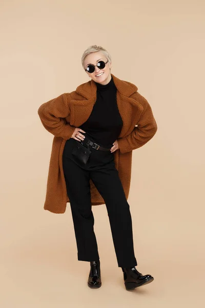 Glad ung hipster kvinna med blont kort hår bär en jacka och solglasögon går över beige bakgrund. — Stockfoto