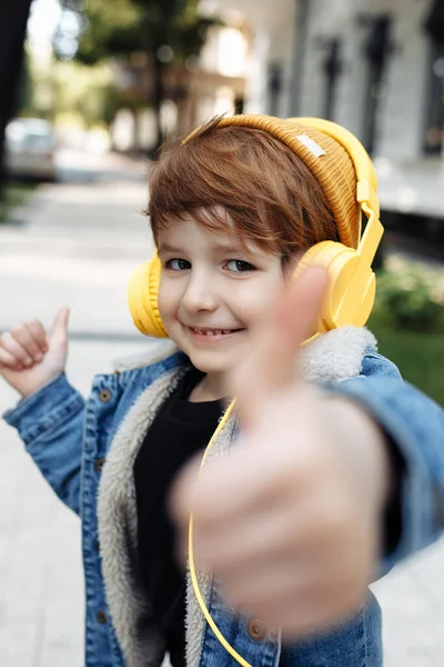 Photo of adorable little boy show thumb up listening music in headphones in street. — Zdjęcie stockowe