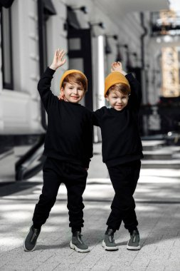 Two cheerful embraced twin boys in casual outfit posing with hands up in the street.