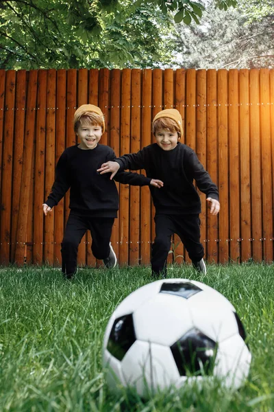 Two cheerful twin boys play soccer with classic ball on the grass at home. — Stockfoto