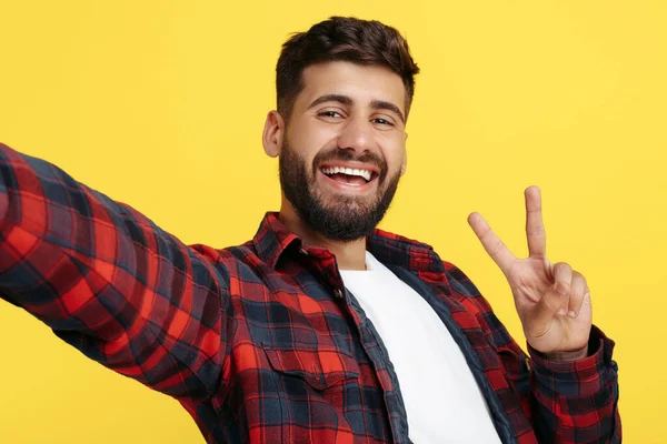 Baard hipster jongeman in geruite shirt selfie met smartphone tonen vrede gebaar over gele achtergrond. — Stockfoto