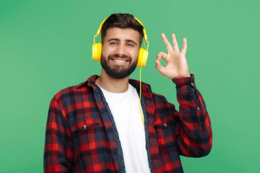 Olumlu sakallı hippi ekose gömlekli, kulaklık takan ve yeşil arka planda iyi niyet gösteren genç adam..