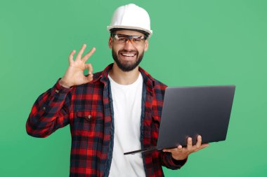 Kendine güvenen sakallı mühendis ya da sıradan giyimli, bilgisayar destekli müteahhit yeşil arka planda iyi niyet gösterisi yapıyor..