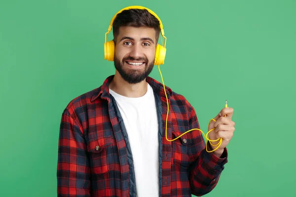 Attrayant hipster barbu jeune homme en chemise à carreaux portant des écouteurs sur fond vert. — Photo