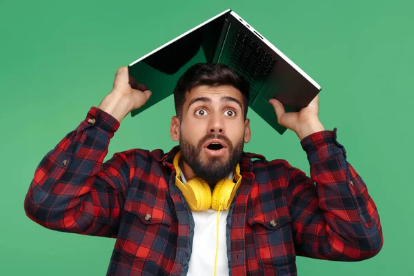 Zszokowany brodaty hipster młody człowiek z komputerem nad głową na zielonym tle. — Zdjęcie stockowe
