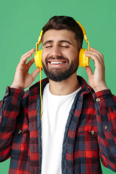 Joven hipster barbudo contento con camisa a cuadros escuchando música en auriculares con los ojos cerrados sobre fondo verde . — Foto de Stock