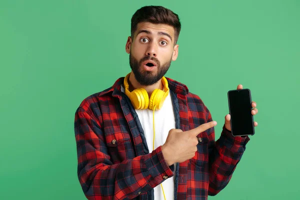 Geschokt bebaarde hipster jongeman met koptelefoon wijzen vinger op smartphone over groene achtergrond. — Stockfoto