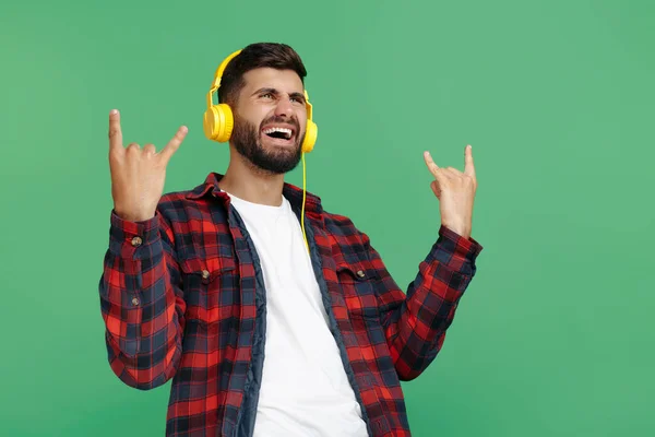 Positieve bebaarde hipster jongeman in geruite shirt met koptelefoon en show rock gebaar over groene achtergrond. — Stockfoto