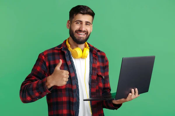 Gelukkig bebaarde hipster jongeman met koptelefoon en computer show duim omhoog over groene achtergrond. — Stockfoto