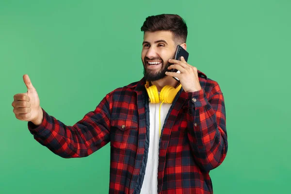 Trendy bebaarde hipster jonge man praten op de smartphone en duim omhoog over groene achtergrond. — Stockfoto
