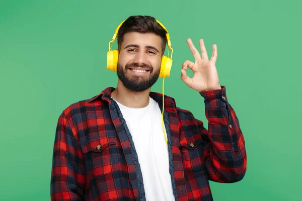 Positieve bebaarde hipster jongeman in geruite shirt met koptelefoon en toon ok gebaar over groene achtergrond. — Stockfoto