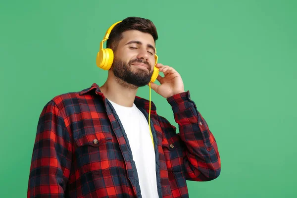 Delizioso giovane hipster barbuto in camicia a quadri che ascolta musica in cuffia con gli occhi chiusi su sfondo verde. — Foto Stock