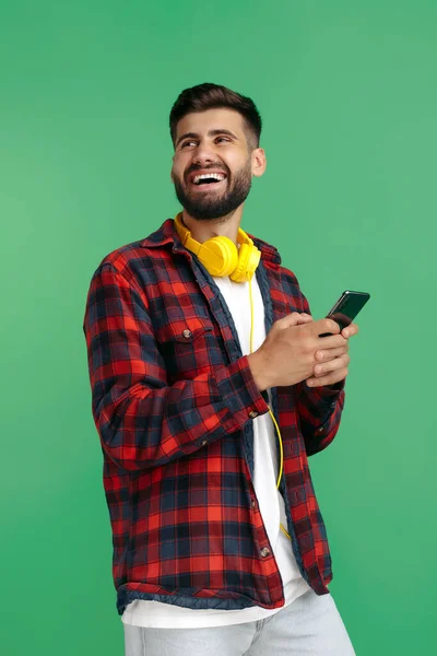 Gelukkig bebaarde hipster jongeman in geruite shirt met koptelefoon gebruik smartphone over groene achtergrond. — Stockfoto