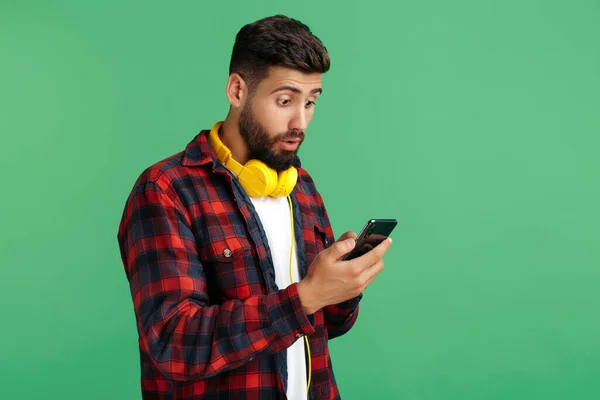 Geschokt bebaarde hipster jongeman met koptelefoon wijzen vinger op smartphone over groene achtergrond. — Stockfoto
