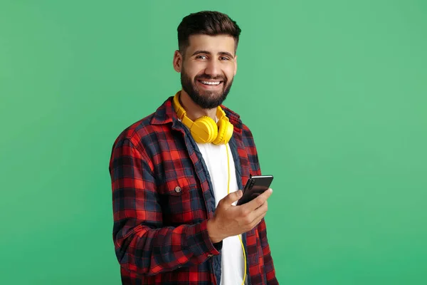 Vrolijke bebaarde hipster jongeman in geruite shirt met koptelefoon gebruik smartphone over groene achtergrond. — Stockfoto