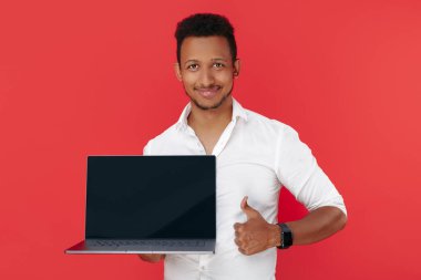 Yakışıklı Afro-Amerikan genç adam dizüstü bilgisayarını tutuyor ve kırmızı arka planda başparmağını gösteriyor..