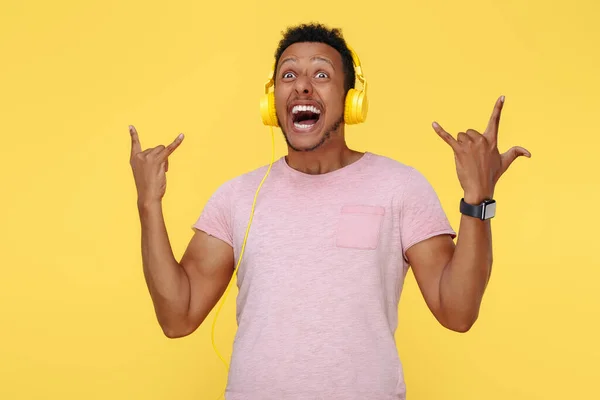 Positieve Afro-Amerikaanse jongeman met een koptelefoon met een rockgebaar over een gele achtergrond. — Stockfoto