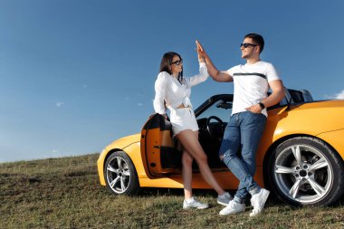 Üstü açık sarı arabanın önünde duran neşeli genç çift. Güzel bir çift beşlik çakıyor..