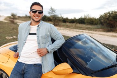 Gülümseyen neşeli genç adam sarı üstü açık arabanın yanında kalıyor. Özgürlük, seyahat ve dikkatsiz konsept.