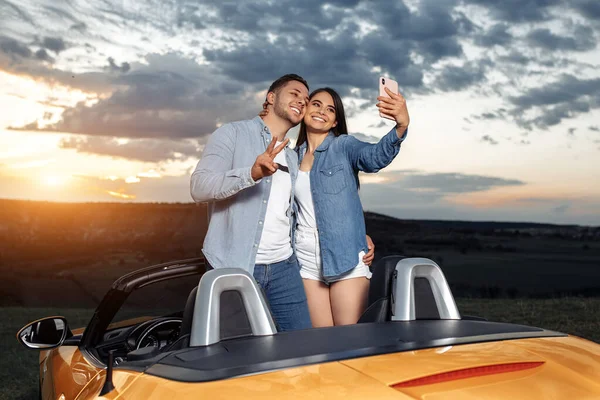 Młoda para jedzie na długą przejażdżkę kabrioletem. Para używać smartfona do selfie. — Zdjęcie stockowe