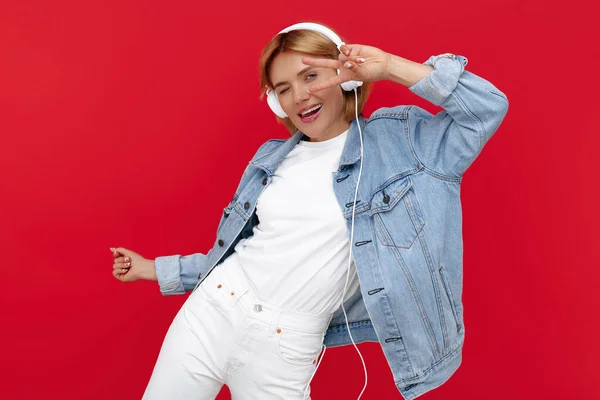 Glänzende junge blonde Frau in lässigem weißen Outfit, die mit Kopfhörern über rotem Hintergrund Musik hört. — Stockfoto