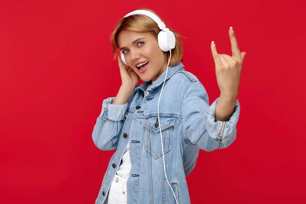 Pozytywna młoda blondynka w luźnym białym stroju słuchająca muzyki w słuchawkach na czerwonym tle. Gest skalny. — Zdjęcie stockowe