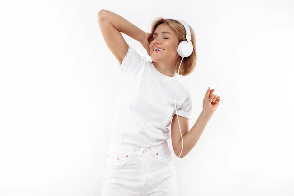 Snygg ung blond kvinna i casual vit outfit lyssna musik i hörlurar över röd bakgrund. — Stockfoto