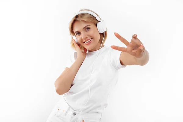 Positive junge blonde Frau in lässigem weißen Outfit, die über Kopfhörer Musik über rotem Hintergrund hört. Friedensgeste. — Stockfoto