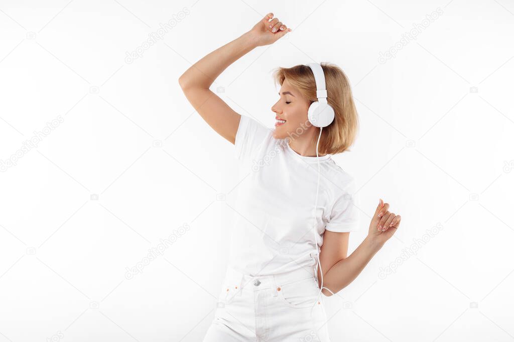 Modern young blonde woman in casual white outfit listening music in headphones over red background.