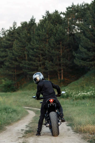 Visão traseira do motociclista sentado na motocicleta ao pôr do sol na estrada do campo . — Fotografia de Stock