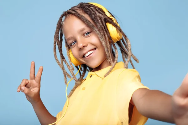 Positiver kleiner Junge mit afrikanischen Ängsten, der Kopfhörer trägt und ein Selfie mit dem Smartphone vor blauem Hintergrund macht. Frieden. — Stockfoto