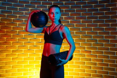 Kararlı genç esmer kadın, neon ışıklarıyla spor salonunda ilaç topu ve köpük jimnastik aletleriyle poz veriyor..
