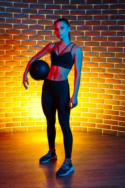 Determined sporty young brunette woman posing with medicine ball in gym in neon lights.