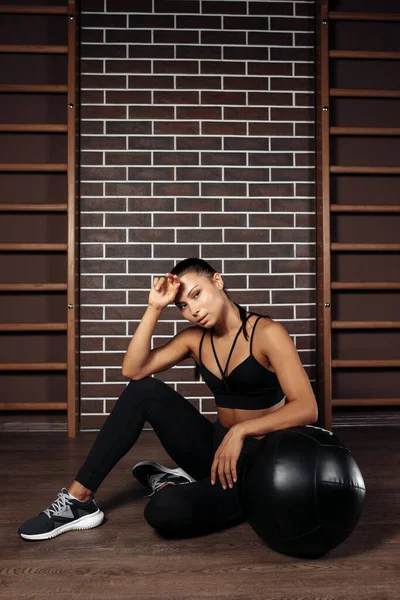 Sporty young brunette woman resting after functional exercises with medicine ball in gym in neon lights. — Stock Photo, Image