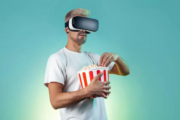 Bearded hipster blonde man wearing virtual reality goggles eating popcorn from bucket in studio in neon lights.