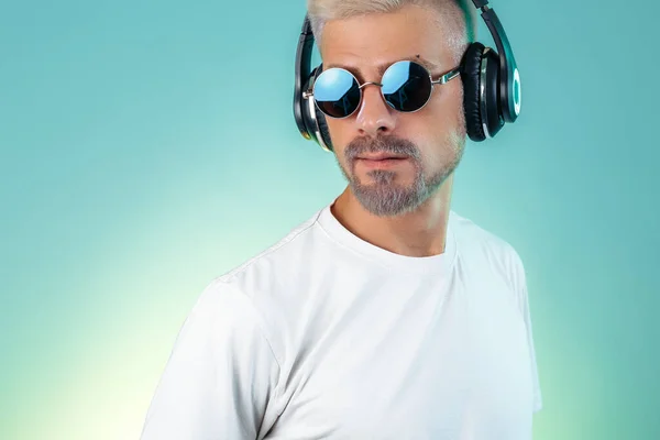 Serieuze bebaarde hippe jongeman met een zonnebril die muziek luistert in stijlvolle koptelefoons op een cyaanse achtergrond. — Stockfoto