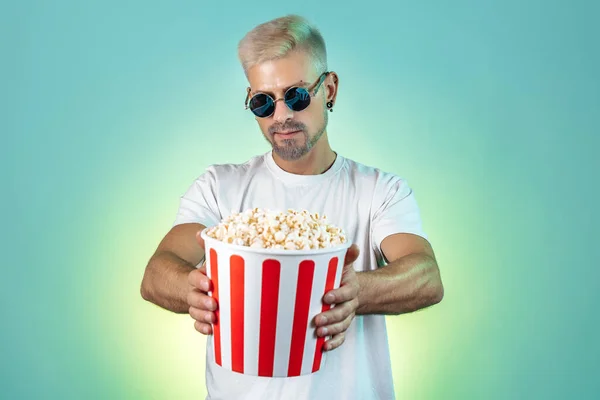 Homme blond hipster barbu portant des lunettes de soleil élégantes et tenant seau avec pop-corn en studio dans des néons. — Photo