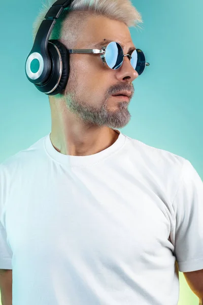 Elegante giovane hipster barbuto che indossa occhiali da sole ascoltare musica in eleganti cuffie su sfondo ciano. — Foto Stock