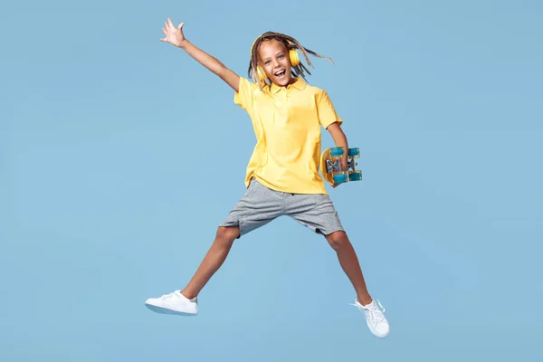 Счастливый маленький мальчик с африканскими дредами в наушниках и скейтборде прыгает через синий фон. — стоковое фото