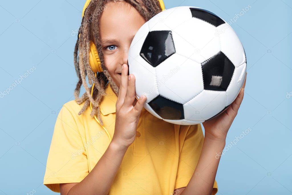 Cheerful little boy with african dreads cover face with soccer ball over blue background.