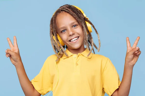 Fröhlicher kleiner Junge mit afrikanischen Ängsten, der über Kopfhörer Musik hört und Friedensgeste vor blauem Hintergrund zeigt. — Stockfoto