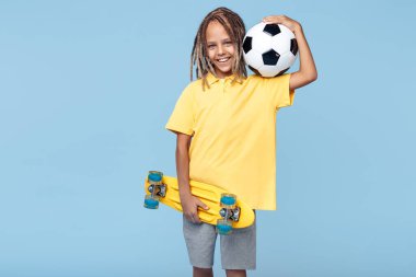 Afrika korkusu olan neşeli küçük çocuk elinde futbol topu ve kaykayı mavi arka planda tutuyor..