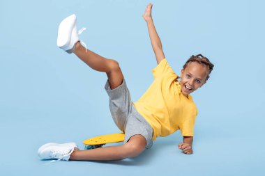 Afrikalı korkusuyla kaykayın üzerinde oturan pozitif küçük çocuk mavi arka planda eğleniyor..