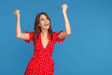 Mutlu genç bir kadın, parlak bir gülümseme içinde, kırmızı bir elbise içinde heyecanlı bir yüzle, mavi arka planda elleri havada yukarı bakıyor..