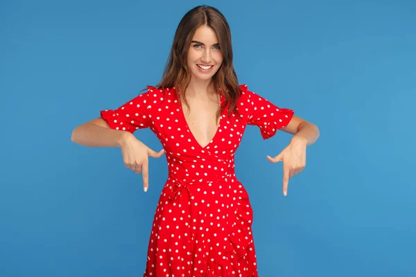 Mujer joven atractiva con sonrisa brillante en vestido rojo señalando los dedos hacia abajo sobre el fondo azul. — Foto de Stock