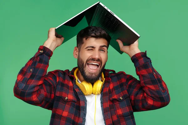 Positieve bebaarde hipster jongeman met computer over hoofd op groene achtergrond. — Stockfoto