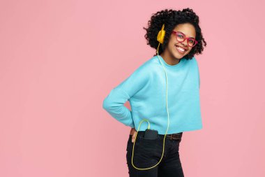 Parlak gülüşlü, pembe arka planda kulaklıkla müzik dinleyen güzel Afrikalı Amerikalı genç bir kadın..