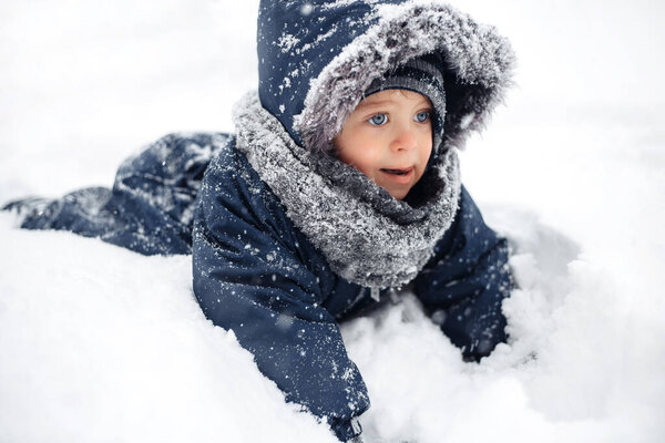 Close-up image of adorable baby boy in a warm white snow suit playing and lying in snow with cute face.